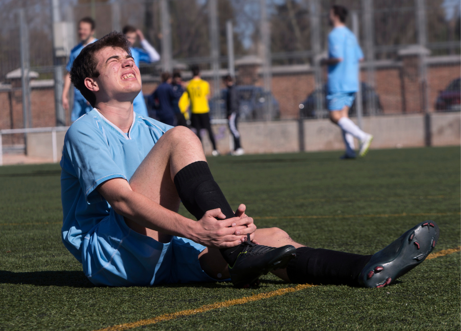 kid on the field that was playing soccer is sitting down holding ankle in pain wearing blue sports uniform
